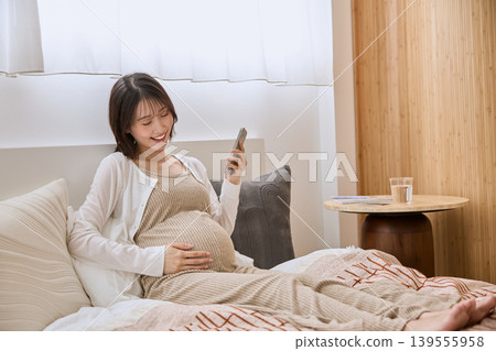一位微笑的孕婦躺在床上,一手放在肚子上,一邊看著她的智慧型手機。 一位微笑的孕婦躺在床上,一手放在肚子上,一邊看著她的智慧型手機。 139555958