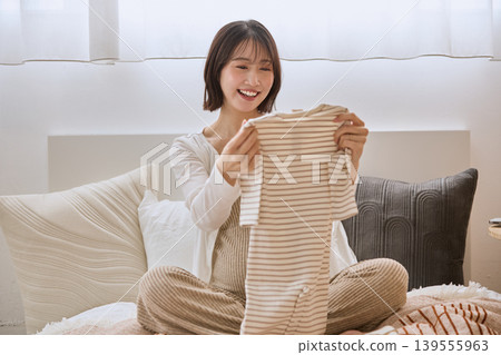 A young woman smiles, holding baby clothes in her hand while lying on a bed. A young woman smiles, holding baby clothes in her hand while lying on a bed. 139555963