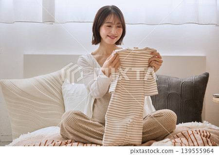 A young woman smiles, holding baby clothes in her hand while lying on a bed. A young woman smiles, holding baby clothes in her hand while lying on a bed. 139555964