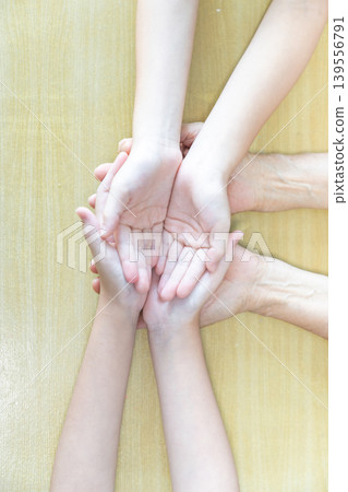 Two children place their hands over an elderly woman hands on a wooden table, creating a close multigeneration family moment of care, support, and trust. Two children place their hands over an elderly woman hands on a wooden table, creating a close multigeneration family moment of care, support, and trust. 139556791