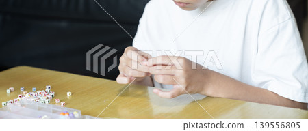A child threads a small colorful bead onto a thin elastic string with both hands. A plastic bead organizer sits blurred in the foreground below. A child threads a small colorful bead onto a thin elastic string with both hands. A plastic bead organizer sits blurred in the foreground below. 139556805