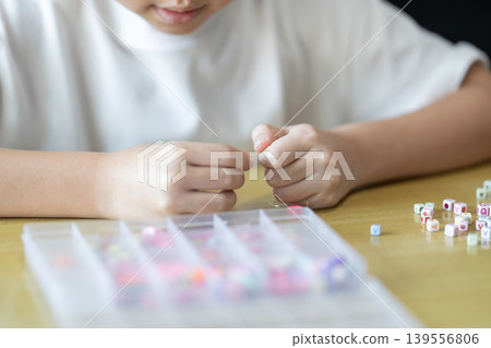 A child threads a small colorful bead onto a thin elastic string with both hands. A plastic bead organizer sits blurred in the foreground below. A child threads a small colorful bead onto a thin elastic string with both hands. A plastic bead organizer sits blurred in the foreground below. 139556806