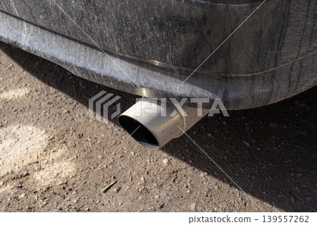 A close view of a car exhaust pipe shows dirt and grime on the outside. The vehicle is parked on a gravel road with sunlight A close view of a car exhaust pipe shows dirt and grime on the outside. The vehicle is parked on a gravel road with sunlight 139557262