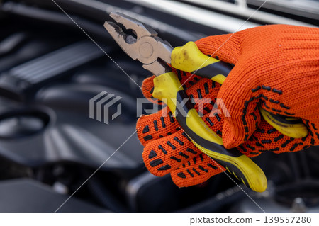 A mechanic holds pliers in one hand while wearing gloves, working on a car engine in a shop during daylight A mechanic holds pliers in one hand while wearing gloves, working on a car engine in a shop during daylight 139557280