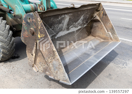 A large tractor bucket is parked on a construction site close to the road. The sun is shining on the equipment A large tractor bucket is parked on a construction site close to the road. The sun is shining on the equipment 139557297