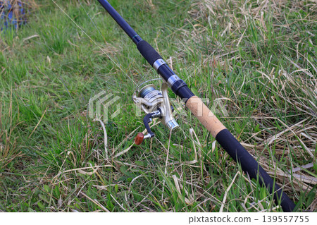 Fishing rod with a golden spinning reel and fishing line in green grass on a riverbank. Concept of outdoor recreation and hobbies. Fishing rod with a golden spinning reel and fishing line in green grass on a riverbank. Concept of outdoor recreation and hobbies. 139557755