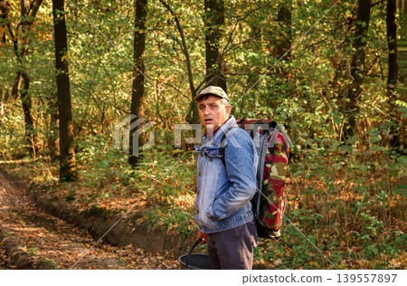 Middle-aged man with a large camouflage backpack and a bucket walking through an autumn forest. Local mushroom picker or hiker in a denim jacket exploring the woods 139557897