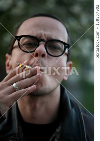 Close-up portrait of a young man with glasses smoking a cigarette. Portrait of a male in a leather jacket exhaling smoke outdoors with a blurred background Close-up portrait of a young man with glasses smoking a cigarette. Portrait of a male in a leather jacket exhaling smoke outdoors with a blurred background 139557902