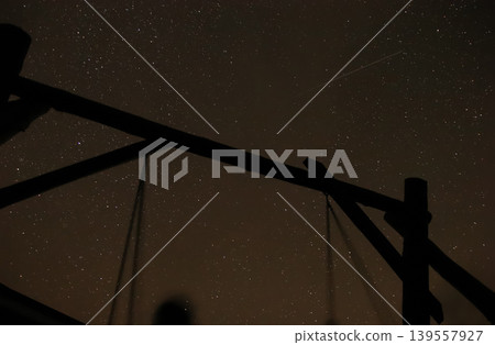 Night sky full of stars with a silhouette of a wooden swing set. Beautiful astrophotography showing constellations and a shooting star or satellite trail in the dark sky Night sky full of stars with a silhouette of a wooden swing set. Beautiful astrophotography showing constellations and a shooting star or satellite trail in the dark sky 139557927