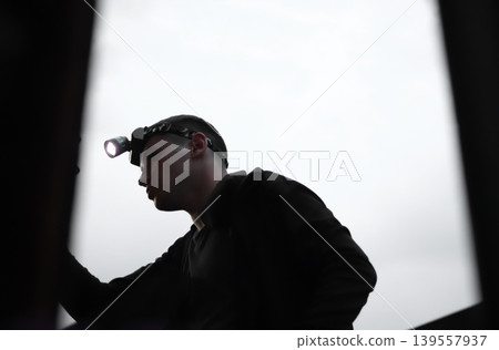 Silhouette of a young man with a headlamp performing a technical inspection or repair. professional lighting equipment. Concept blackout. Ukraine 139557937