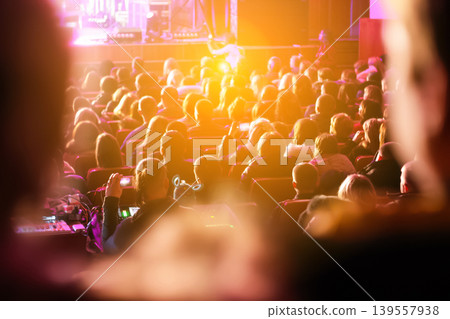 Crowd of people sitting in a theater or concert hall during a performance. Rear view of an audience in a dimly lit auditorium with bright stage lights and warm atmosphere 139557938