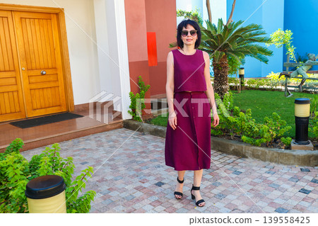 A woman who owns a large house stands near the front door against the backdrop of a green garden A woman who owns a large house stands near the front door against the backdrop of a green garden 139558425