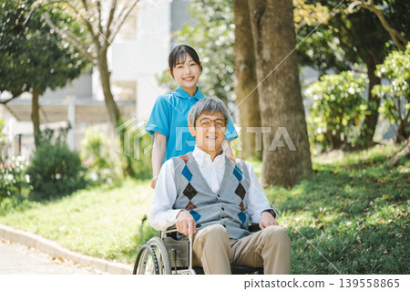 A young caregiver and a senior man strolling through a park in a wheelchair. 139558865