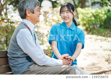一位年輕的護理員坐在公園長椅上，旁邊有一位老人。 139558870