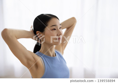 A middle-aged Japanese woman in yoga wear ties her hair up by a bright window. A middle-aged Japanese woman in yoga wear ties her hair up by a bright window. 139558894