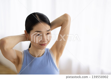 一位身穿瑜珈服的中年日本女性在明亮的窗邊紮起頭髮。 139558902