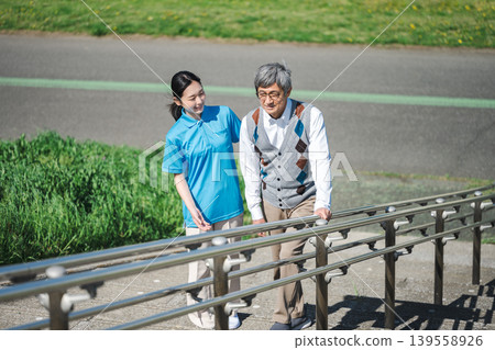 A senior man climbing the stairs in a park, accompanied by a female caregiver assisting him. A senior man climbing the stairs in a park, accompanied by a female caregiver assisting him. 139558926