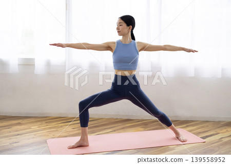 A middle-aged Japanese woman performs the warrior pose (yoga) on a mat by a bright window. A middle-aged Japanese woman performs the warrior pose (yoga) on a mat by a bright window. 139558952