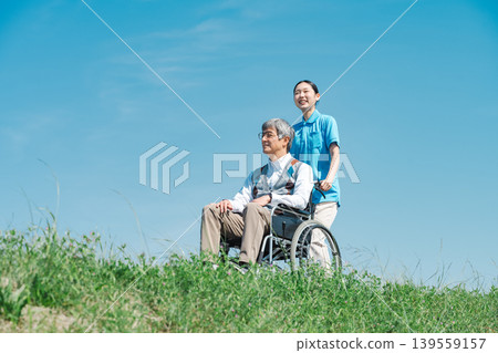 A caregiver and a senior man strolling through a park in a wheelchair. 139559157