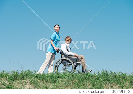 A caregiver and a senior man strolling along the riverbank in a wheelchair. A caregiver and a senior man strolling along the riverbank in a wheelchair. 139559160