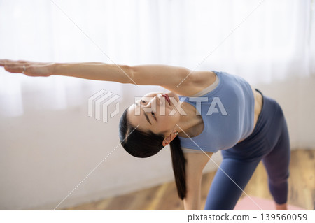 一位日本女子在明亮的窗戶旁的墊子上練習半月式（Ardha Chandrasana）。 139560059
