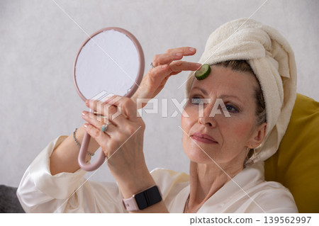 Woman applying cucumber slice on forehead while looking in mirror at home during self-care routine Woman applying cucumber slice on forehead while looking in mirror at home during self-care routine 139562997