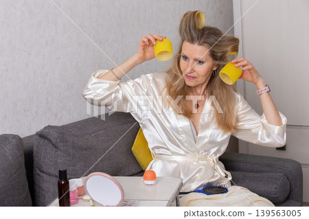 Woman styles her hair with curlers in a cozy living room setting during morning routine Woman styles her hair with curlers in a cozy living room setting during morning routine 139563005