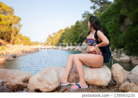 A woman sits on a rock by the river, wearing a bikini and carrying a backpack in the mountains. She looks at the surrounding nature. A woman sits on a rock by the river, wearing a bikini and carrying a backpack in the mountains. She looks at the surrounding nature. 139563220