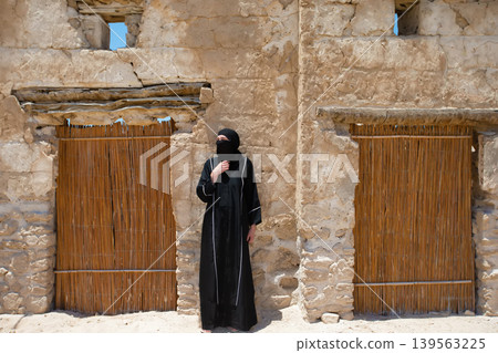 Woman in traditional attire stands beside rustic adobe structure in desert at midday Woman in traditional attire stands beside rustic adobe structure in desert at midday 139563225