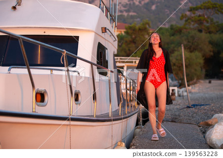 Woman enjoying sunset stroll along dock in red swimsuit and cover-up near moored boat by the shore 139563228