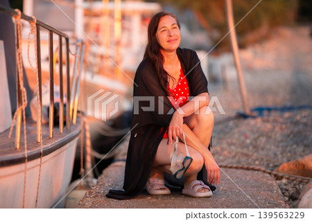 Smiling woman in a red dress enjoying sunset by the marina in coastal town Smiling woman in a red dress enjoying sunset by the marina in coastal town 139563229