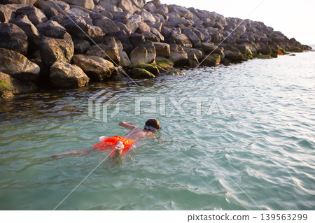 Child swims near rocky shoreline at sunset in tranquil coastal scene Child swims near rocky shoreline at sunset in tranquil coastal scene 139563299