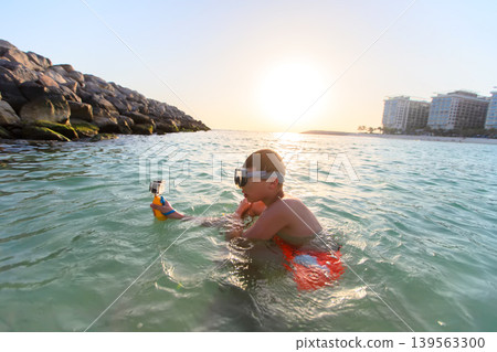 A young boy is immersed in shallow water, focused on his camera as he captures the beauty of the underwater world. The sun sets on the horizon, casting a warm glow. A young boy is immersed in shallow water, focused on his camera as he captures the beauty of the underwater world. The sun sets on the horizon, casting a warm glow. 139563300