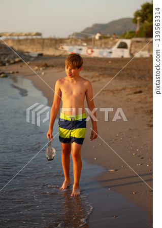 The boy walks on the beach at sunset, holding a mask next to calm waves and a rocky shore 139563314