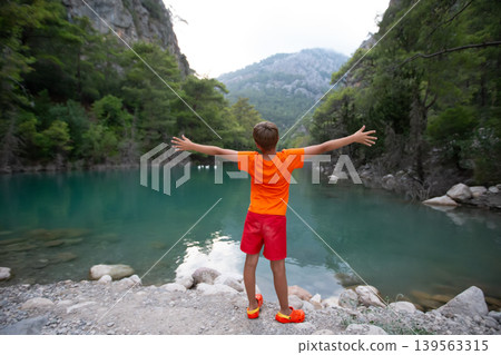 Child enjoys nature by the serene lake surrounded by mountains and trees during a peaceful summer day Child enjoys nature by the serene lake surrounded by mountains and trees during a peaceful summer day 139563315