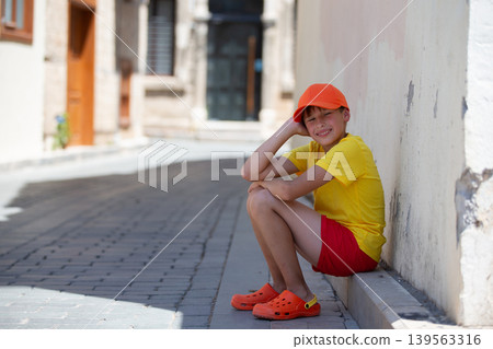 Boy enjoying a sunny day in a narrow street while wearing bright clothing and sitting on the curb Boy enjoying a sunny day in a narrow street while wearing bright clothing and sitting on the curb 139563316