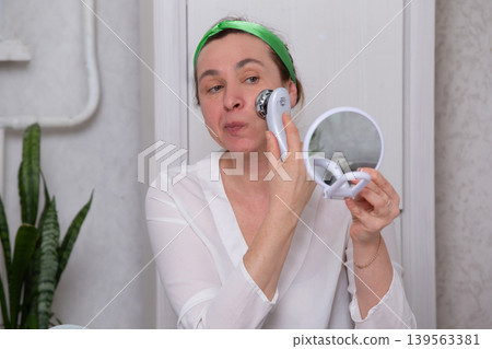 Woman uses facial cleansing device while looking in mirror at home during skincare routine 139563381