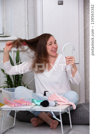 Woman grooming her hair while sitting on a comfortable surface in a well-lit indoor setting with hair tools around her 139563382