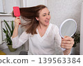 Woman brushing her hair while looking in a mirror at home during daylight hours 139563384