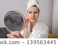 An elderly woman uses a skin care product, looking in the hand of a mirror at home in the daytime 139563445