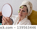 An elderly woman enjoying a relaxing routine on the skin care of the house, looking in the mirror with slices of cucumber on the forehead 139563446