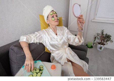 Elderly woman relaxes with pieces of cucumber on her face, looking in the mirror at home during self -service Elderly woman relaxes with pieces of cucumber on her face, looking in the mirror at home during self -service 139563447
