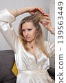 Woman styling her hair while wearing a silk robe in a cozy indoor setting during the afternoon 139563449