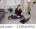 Woman working from home in a cozy space with a cat nearby while sitting on a mat during daylight hours 139563452