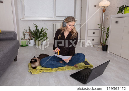Woman working from home in a cozy space with a cat nearby while sitting on a mat during daylight hours Woman working from home in a cozy space with a cat nearby while sitting on a mat during daylight hours 139563452