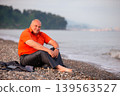Man in orange shirt relaxes by the pebbled beach in the early morning light with waves gently lapping at the shore 139563527