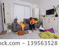 A teenager sits on the couch in a dirty living room, focused on the phone among the mess and colorful decor 139563528