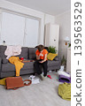 A disorganized living space shows a teenager lost in thought, sitting on a sofa with a scattered disorder around 139563529