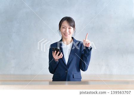 一名身穿西裝的女子一邊指著什麼,一邊拿著智慧型手機。 一名身穿西裝的女子一邊指著什麼,一邊拿著智慧型手機。 139563707