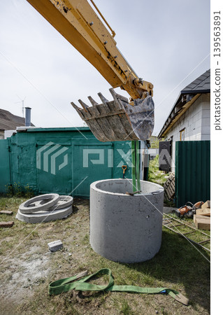 A machine works to place a green pipe inside a concrete well. The scene shows a construction site in a residential area with buildings in the background A machine works to place a green pipe inside a concrete well. The scene shows a construction site in a residential area with buildings in the background 139563891